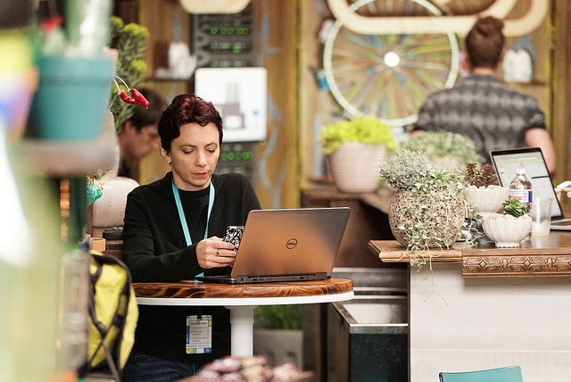 a woman named Laura Pevehouse stands in front of a laptop looking at her cell phone wearing the lanyard of an event she is attending.